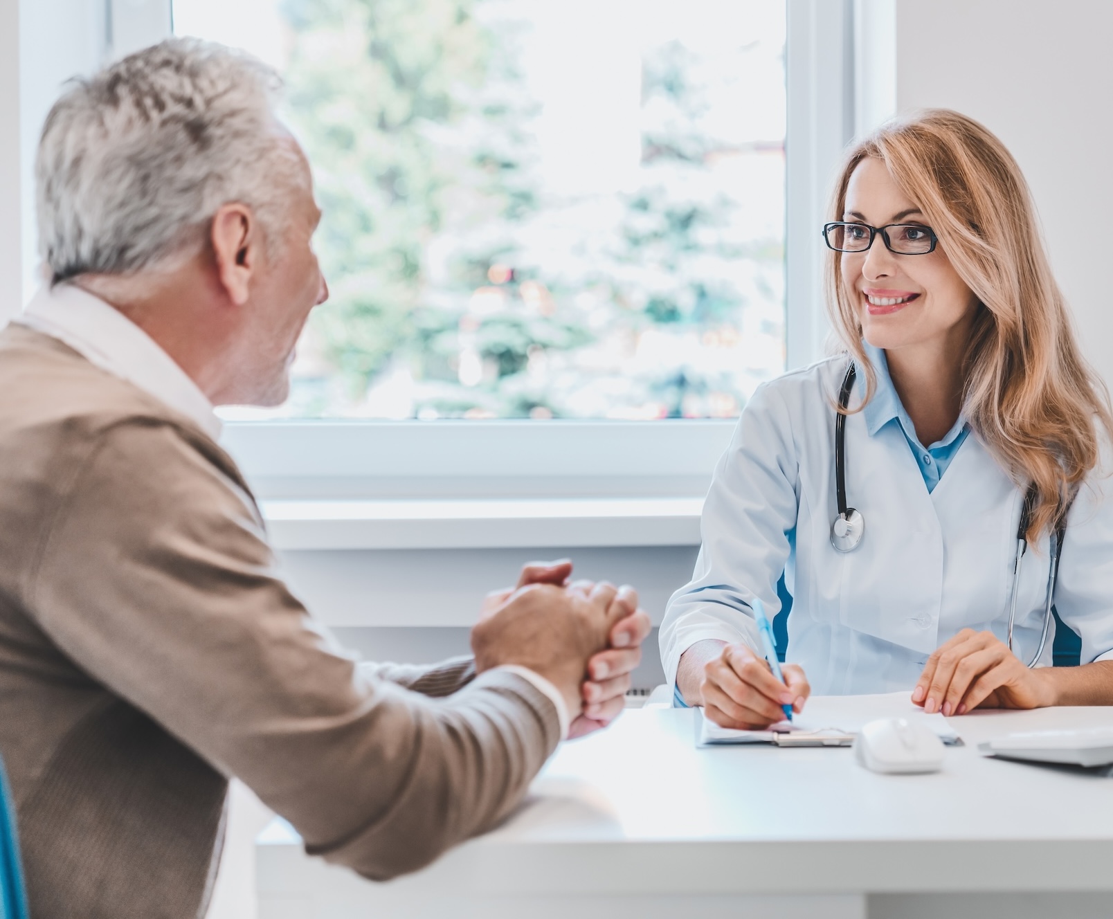 Smiling female doctor with glasses and stethoscope talking to an older male patient at a bright medical office.